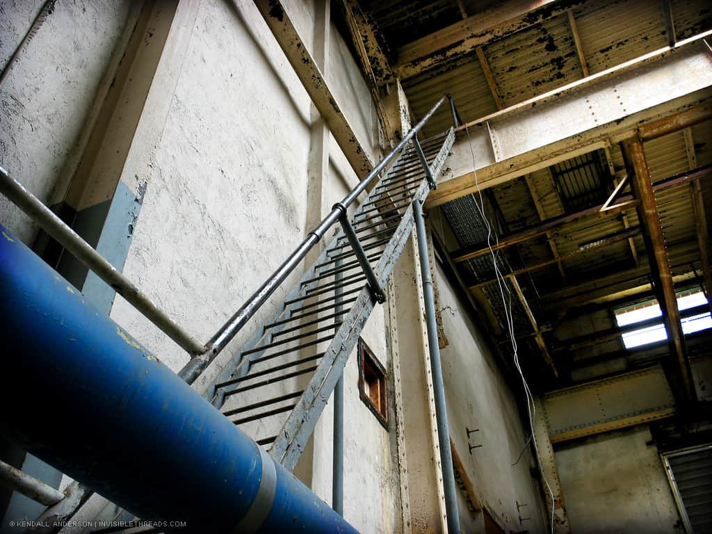 A steep ladder of bars (rather than steps) extends up 3 storeys to the ceiling attic space in a mechanical room.