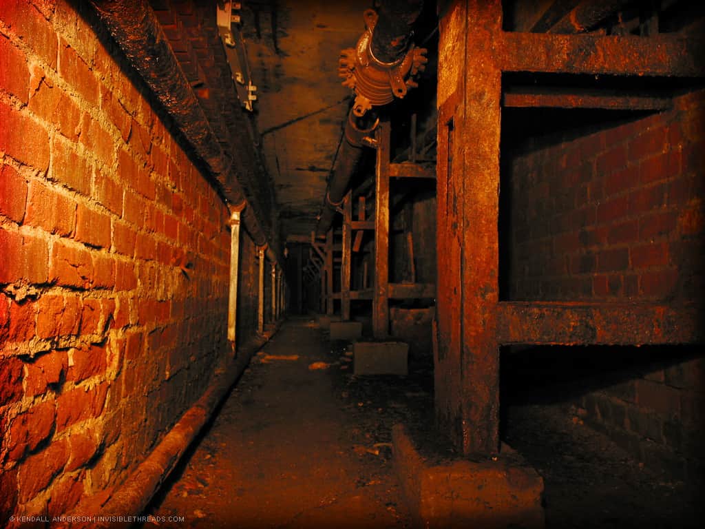 An underground small steam tunnel is lined with red brick walls. Pipes run along the ceiling.