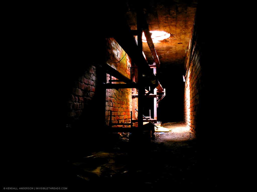 A dark tunnel lined with red bricks has a hole in the ceiling where light enters. Pipes run along the ceiling.