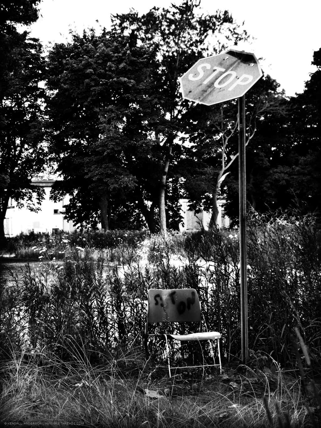 A chair is on the ground outside, beside a broken vehicle Stop sign. Someone has written the words 'sit on' on the back of the chair.