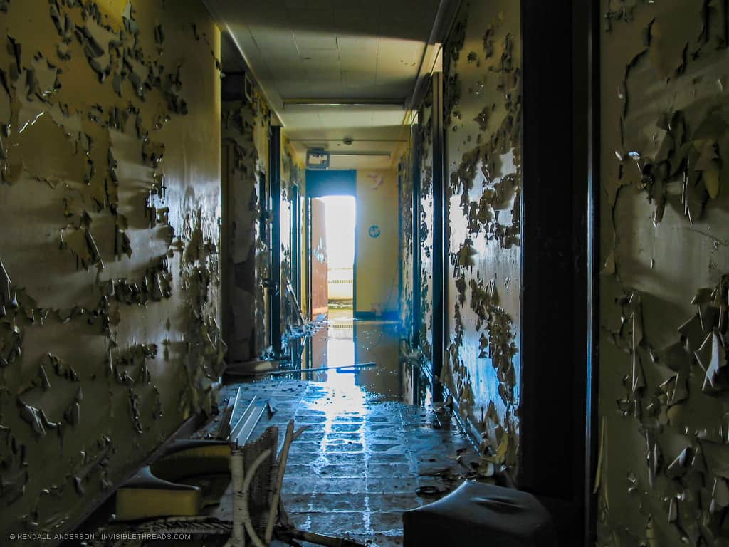 A dark interior corridor leads to a bright window. The hallway is flooded with water and all walls have large amounts of paint peeling off them.