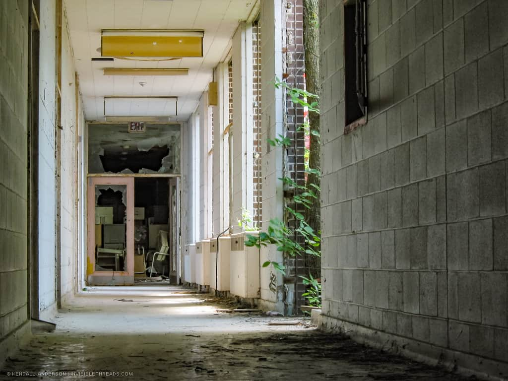 A bright interior corridor of concrete block has windows to a courtyard. All windows and the exit door contain broken glass.