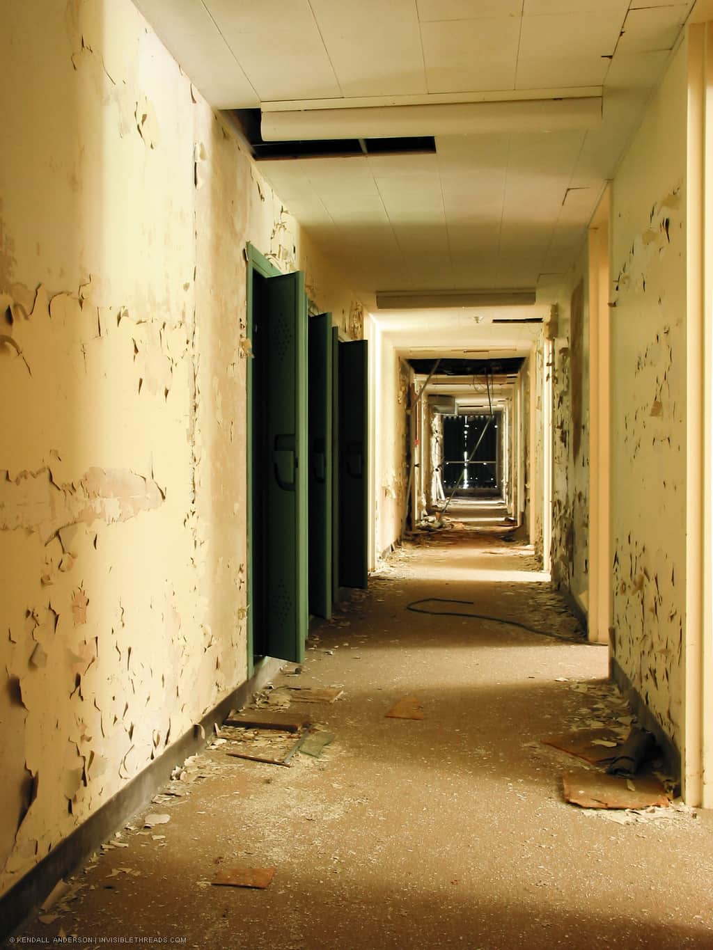 A carpeted hallway with yellow walls and some green lockers is covered with peeling paint and general debris.