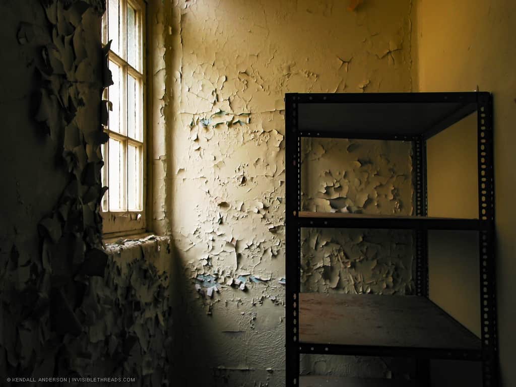 A small closet with a metal shelving unit is lit by a corner window. The walls are covered with cracking and peeling paint.