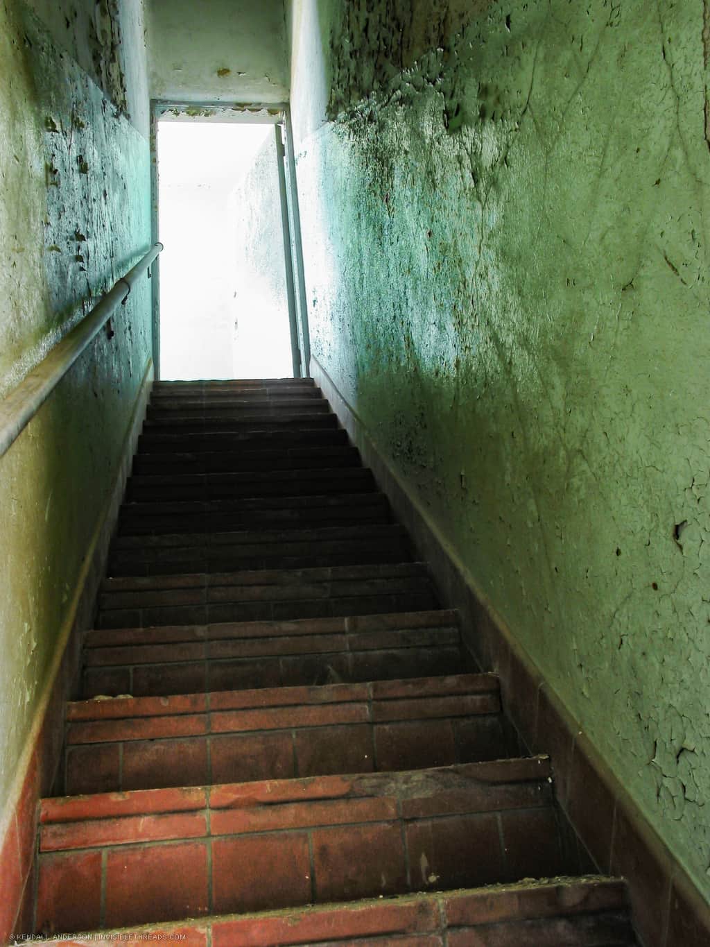 A stair leads up to the second floor, which is bright. The walls beside the stairs are green, dirty, and cracked.