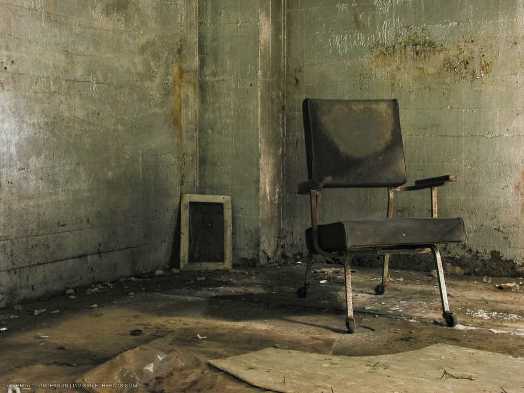 A grey office chair sits in the corner of an empty room. The surrounding concrete walls are dirty and grimy.