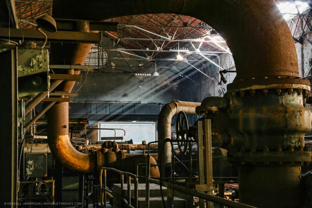 Two large rusting pipes frame a view of the industrial building interior. The pipes have an orange glow from the sunlight streaming in from the skylights above. The rest of the building is densely packed with pipes and metal railings.