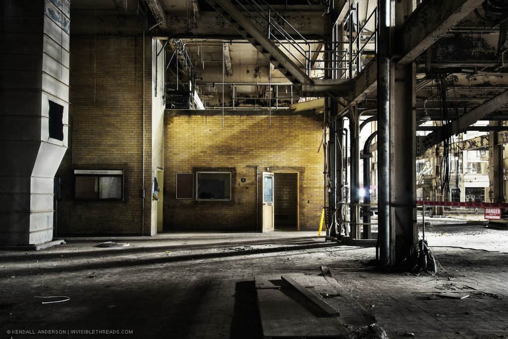 A yellow brick wall has an opening with a door and a small window. The wall is inside a large industrial power station, surrounded by exposed steel columns and beams. A large metal duct rises on the left and goes up above the edge of the image. Debris is seen around the floor.