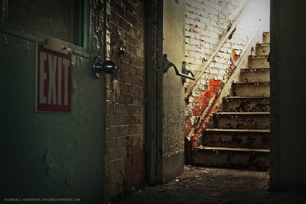 An open door, at the bottom of a dirty staircase with a peeling, painted brick wall.