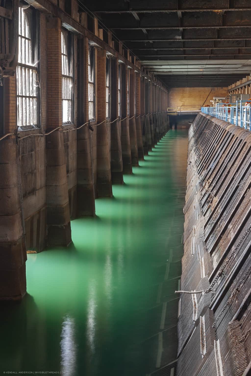 The length of the forebay is full of bright green water. On the left is the exterior stone wall with regular columns and windows to the outside. On the right are the angled steel grates that prevent ice and trash from entering the penstocks.