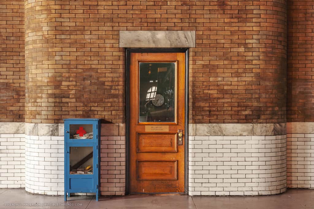 A wooden door with a large window is in the middle of a brick wall inside a building. The brick is white until it reaches the height of the door handle. Above, the brick is reddish-orange. A blue first aid cabinet stands to the left of the door.