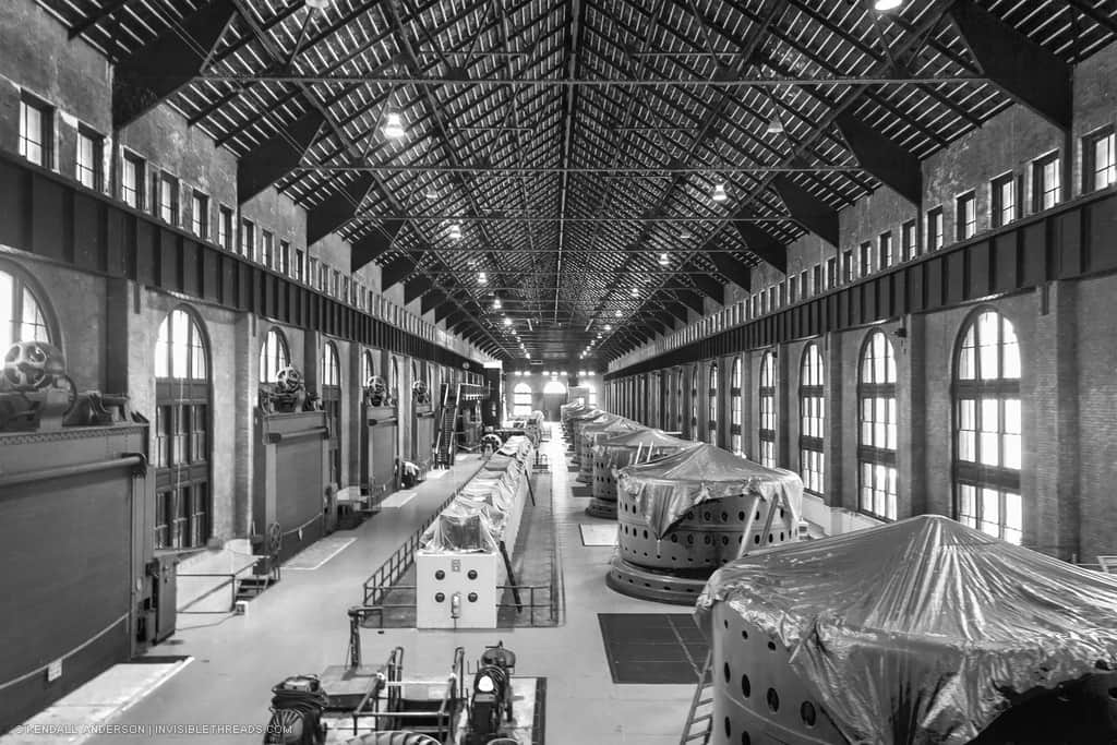 The entire turbine generator hall of the power station is shown in perspective. The generator heads are lined up on the right, covered in tarps. The interior is brightly lit by the large windows in the brick wall.