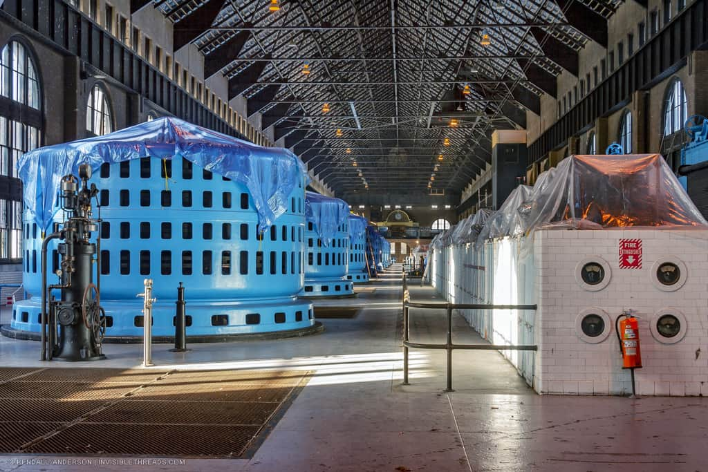 The central corridor down the main turbine generator hall of the power station is lined with blue generator heads (the alternators) on the left, and a bank of equipment surrounded in white brick (the bus bar) on the right.