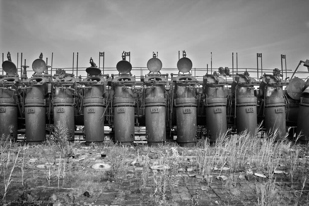 A row of cylinders is lined up on top of an industrial coke battery. Each cylinder is the elbow, or standpipe cover, for the pipe below. The entire set is overgrown and abandoned.