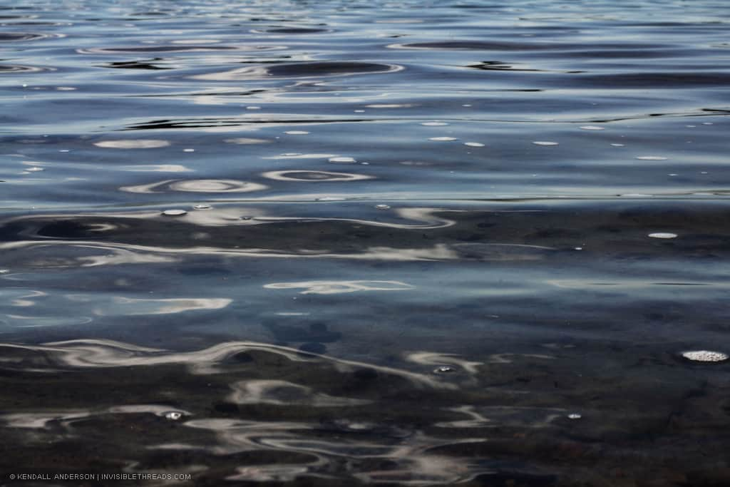 Blue lake water, with light and dark spots and some visibility into the shallow lake bottom below.