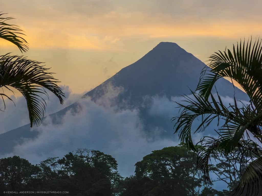 Volcano peak silhouetted against the light yellow sky at dawn. Jungle trees and palm leaves frame the volcano.