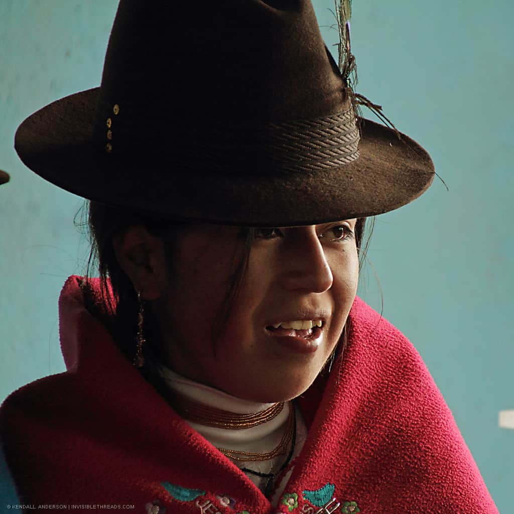 Portrait of a young indigenous girl, smiling and wearing traditional clothes: a bright red shawl and a brown fedora hat.