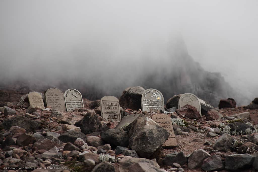 A group of 7 tombstones are set in the rocky mountain hillside, with thick fog and mist behind, obscuring Mount Chimborazo behind in the distance.