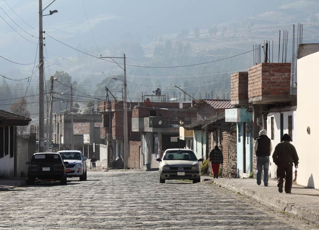 Rural town streetscape early in the morning, with people on sidewalk beside cobblestone streets. Hazy tree-covered hillside in the distance.