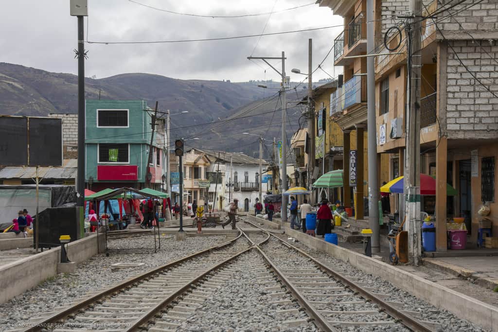 Two train tracks connect into one, running between buildings. Market shops and storefronts are adjacent to the tracks, with people selling things.