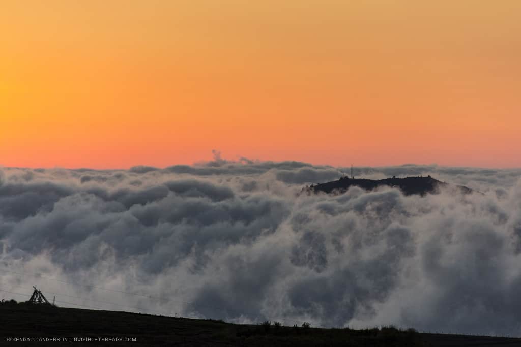 A silhouetted hill top rises above the clouds with a bright orange sunset sky behind.