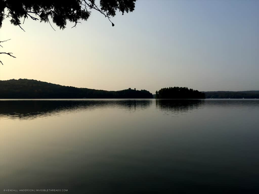 Calm lake water in the early morning at dawn, with the silhouette of an island and the treeline in the distance on the horizon.