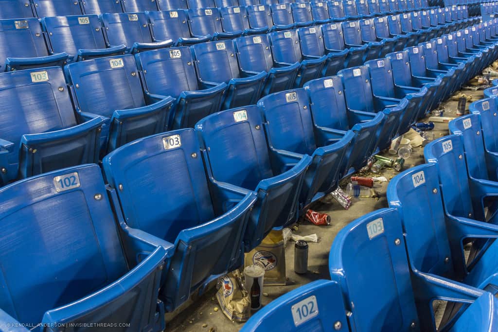 Rows of blue empty baseball bleachers, with discarded bags, bottles, cans and garbage on the ground below
