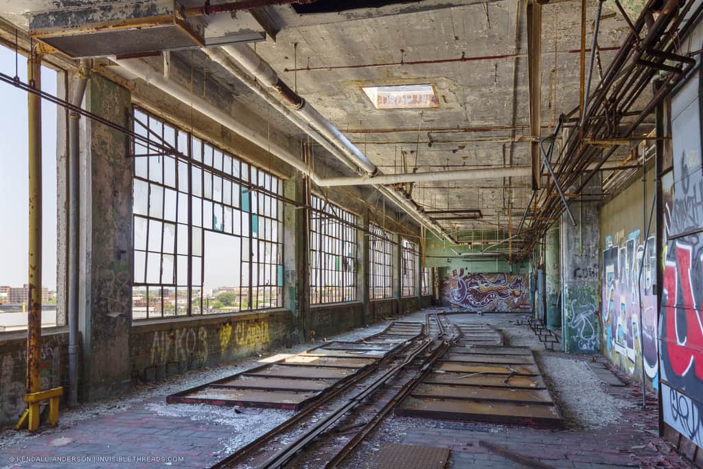 Rails on the floor inside an abandoned factory warehouse with graffiti, with windows looking to the city of Detroit
