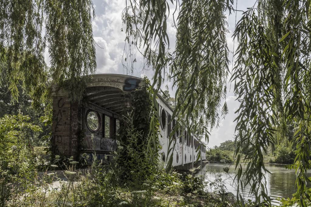 An abandoned subway train car, functioning as a bridge over a river, overgrown with vegetation