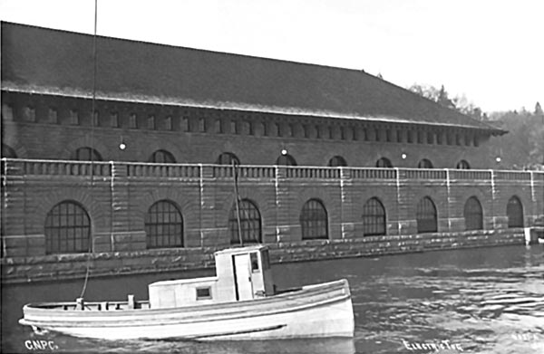 A small boat is in the water (the exterior forebay) in front of the exterior wall of Rankine Generating Station. Archival photo from 1906.
