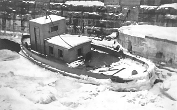 An ice breaker tug boat is surrounded by thick ice in the water, pressed up against the exterior wall of the Rankine Generating Station. Archival photo from 1936.