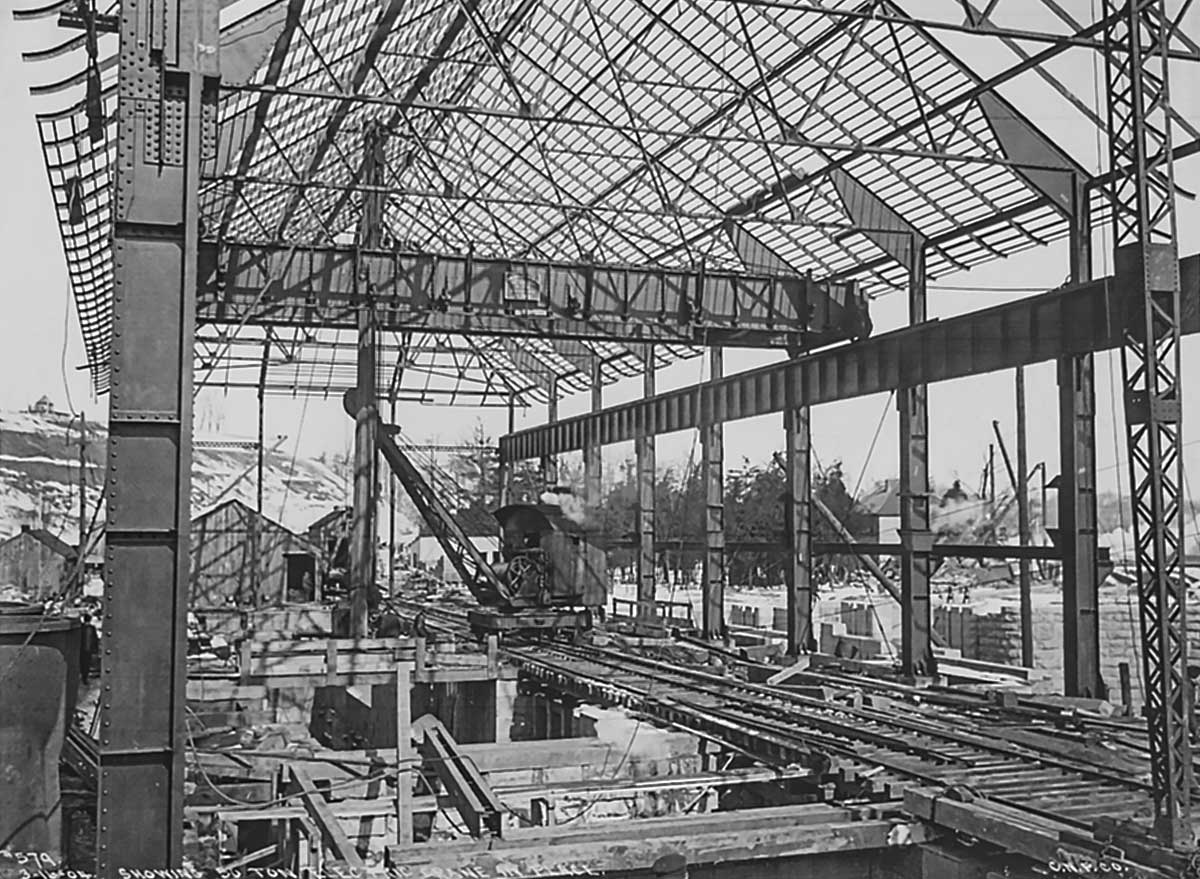 Archival photo of Rankine Generating Station power house under construction, from 1904. Two sections of steel columns are standing, along with the roof trusses connecting them. A small crane on railway tracks inside the frame is moving materials around.